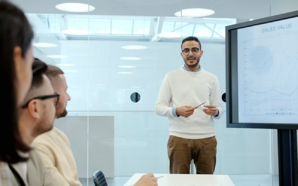 Man pointing at a bar chart on a wall-mounted screen labeled Sales Value in an office